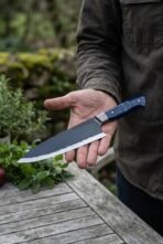 Open palm holding a high carbon chef knife with blue resin handle and Damascus bolster over an outdoor wooden table