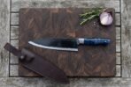 Top view of a black finished chef knife with blue resin handle on an end grain cutting board next to its brown leather sheath