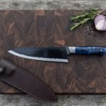 Top view of a black finished chef knife with blue resin handle on an end grain cutting board next to its brown leather sheath