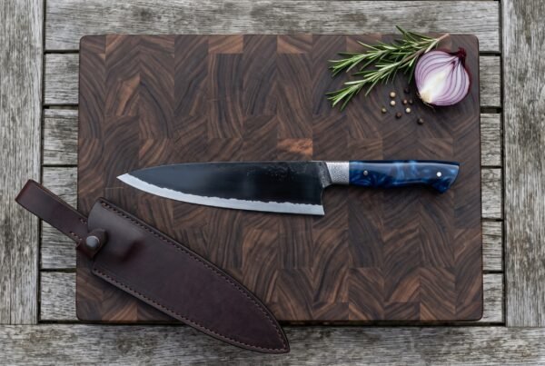 Top view of a black finished chef knife with blue resin handle on an end grain cutting board next to its brown leather sheath
