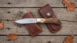 Open stag horn folding pocket knife laid over two brown leather sheaths on weathered planks with oak leaves
