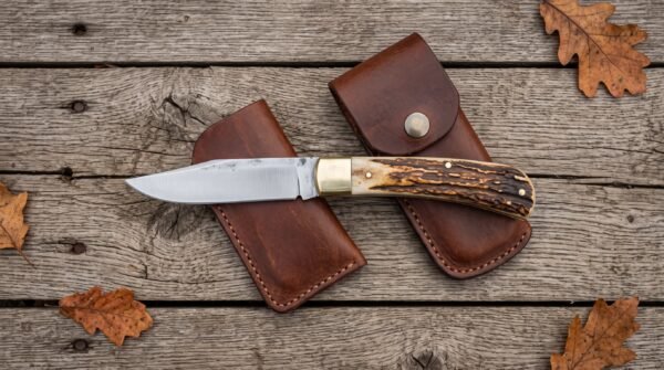 Open stag horn folding pocket knife laid over two brown leather sheaths on weathered planks with oak leaves