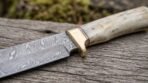 Damascus steel seax knife with pale bone handle and brass bolster, close up of the flowing blade pattern on weathered wood