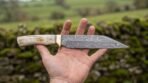 Damascus steel seax knife with natural bone handle held flat across an open palm in front of a mossy stone wall