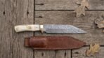 Damascus steel seax knife with bone handle and brass bolster above its stitched brown leather sheath on planks with oak leaves