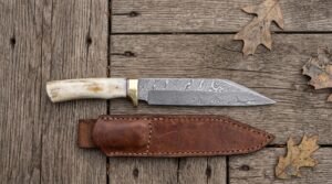 Damascus steel seax knife with bone handle and brass bolster above its stitched brown leather sheath on planks with oak leaves