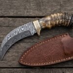 Damascus steel hawkbill knife with ram horn handle and brass bolster laid beside its brown leather sheath on dark planks