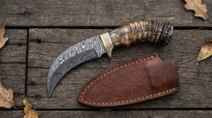 Damascus steel hawkbill knife with ram horn handle and brass bolster laid beside its brown leather sheath on dark planks