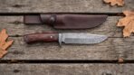 Damascus steel camp knife with rosewood handle and steel guard laid flat on weathered planks under its brown leather sheath