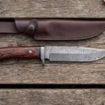 Damascus steel camp knife with rosewood handle and steel guard laid flat on weathered planks under its brown leather sheath