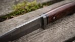 Close up of a Damascus steel camp knife blade with steel guard, black spacer and dark rosewood handle on old grey wood