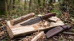 Damascus steel camp knife with rosewood handle resting on a split log among wood shavings beside a brown leather sheath