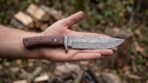 Damascus steel camp knife with rosewood handle and pin lying across an open palm in a woodland clearing with stacked logs
