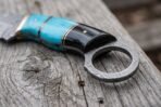 Close up of a Damascus karambit handle in turquoise and black horn with brass spacers and a Damascus finger ring on old wood