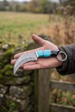 Curved Damascus karambit with turquoise and black handle held flat on an open hand by a wooden gate and stone wall