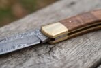 Damascus folding pocket knife in close view at the brass bolster, walnut wood handle and patterned blade on old wood