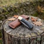 Damascus folding pocket knife closed on a tree stump with black buffalo horn handle, brass bolster and leather sheath