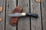 Damascus folding pocket knife lying open across a tan leather sheath on grey weathered decking with autumn leaves