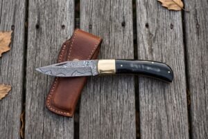 Damascus folding pocket knife lying open across a tan leather sheath on grey weathered decking with autumn leaves