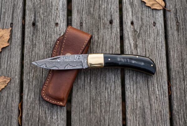 Damascus folding pocket knife lying open across a tan leather sheath on grey weathered decking with autumn leaves