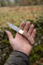 Damascus folding pocket knife with walnut wood handle and brass bolster held open on a palm over an autumn field