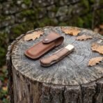 Damascus folding pocket knife with walnut wood handle shown closed on a tree stump beside its tan leather sheath