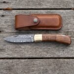 Damascus folding pocket knife with walnut wood handle lying open on grey boards from above, leather sheath above it