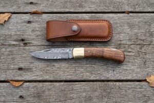 Damascus folding pocket knife with walnut wood handle lying open on grey boards from above, leather sheath above it