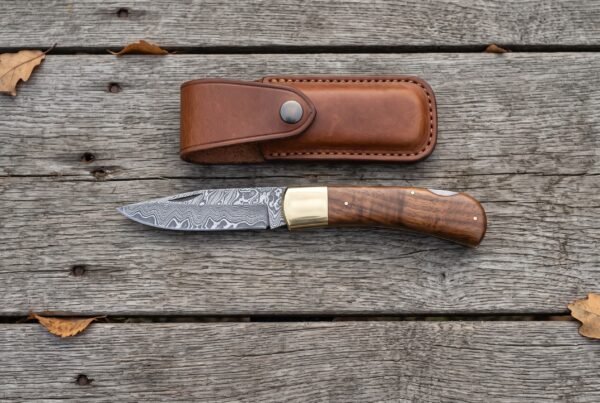 Damascus folding pocket knife with walnut wood handle lying open on grey boards from above, leather sheath above it