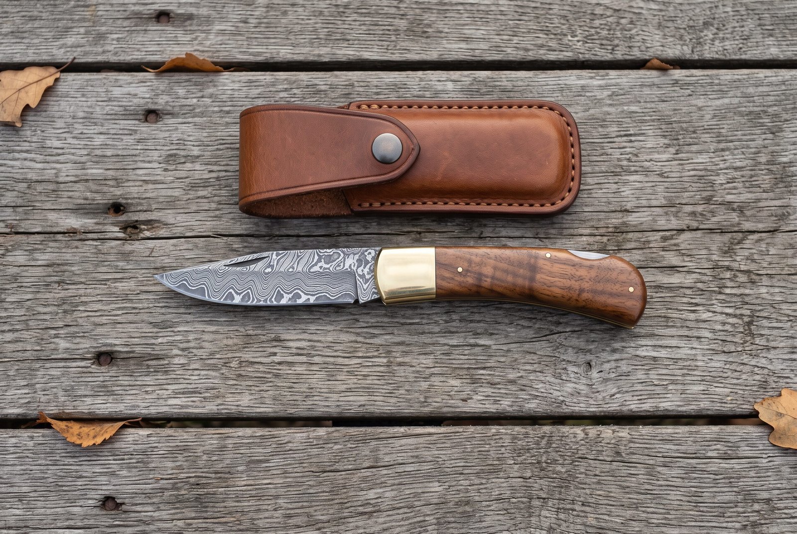 Damascus folding pocket knife with walnut wood handle lying open on grey boards from above, leather sheath above it Damascus folding pocket knife with walnut wood handle lying open on grey boards from above, leather sheath above it