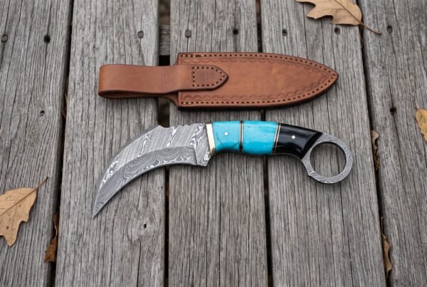 Damascus karambit knife with finger ring and turquoise and black horn handle laid below its tan leather sheath on wooden boards