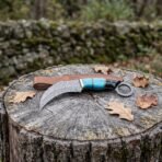 Damascus karambit knife with turquoise and black horn handle and finger ring on a tree stump beside its brown leather sheath