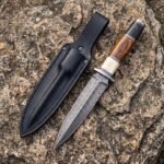 Damascus steel dagger with bone, wood and black handle spacers beside a black leather sheath on lichen covered stone