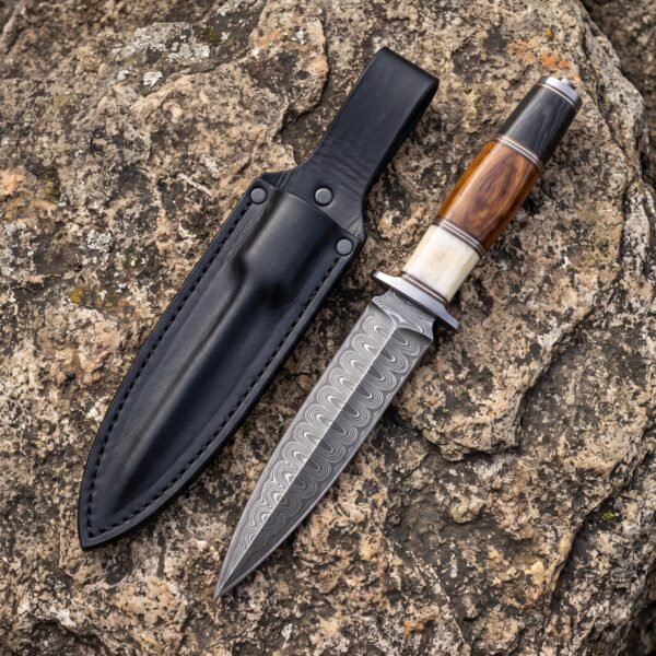 Damascus steel dagger with bone, wood and black handle spacers beside a black leather sheath on lichen covered stone