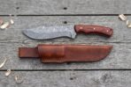 Damascus steel tracker knife with rosewood handle from above on grey boards, brown leather sheath laid below it