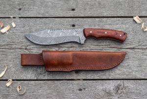 Damascus steel tracker knife with rosewood handle from above on grey boards, brown leather sheath laid below it