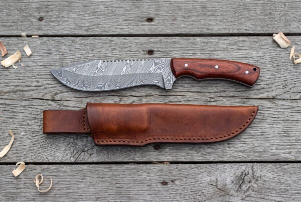 Damascus steel tracker knife with rosewood handle from above on grey boards, brown leather sheath laid below it