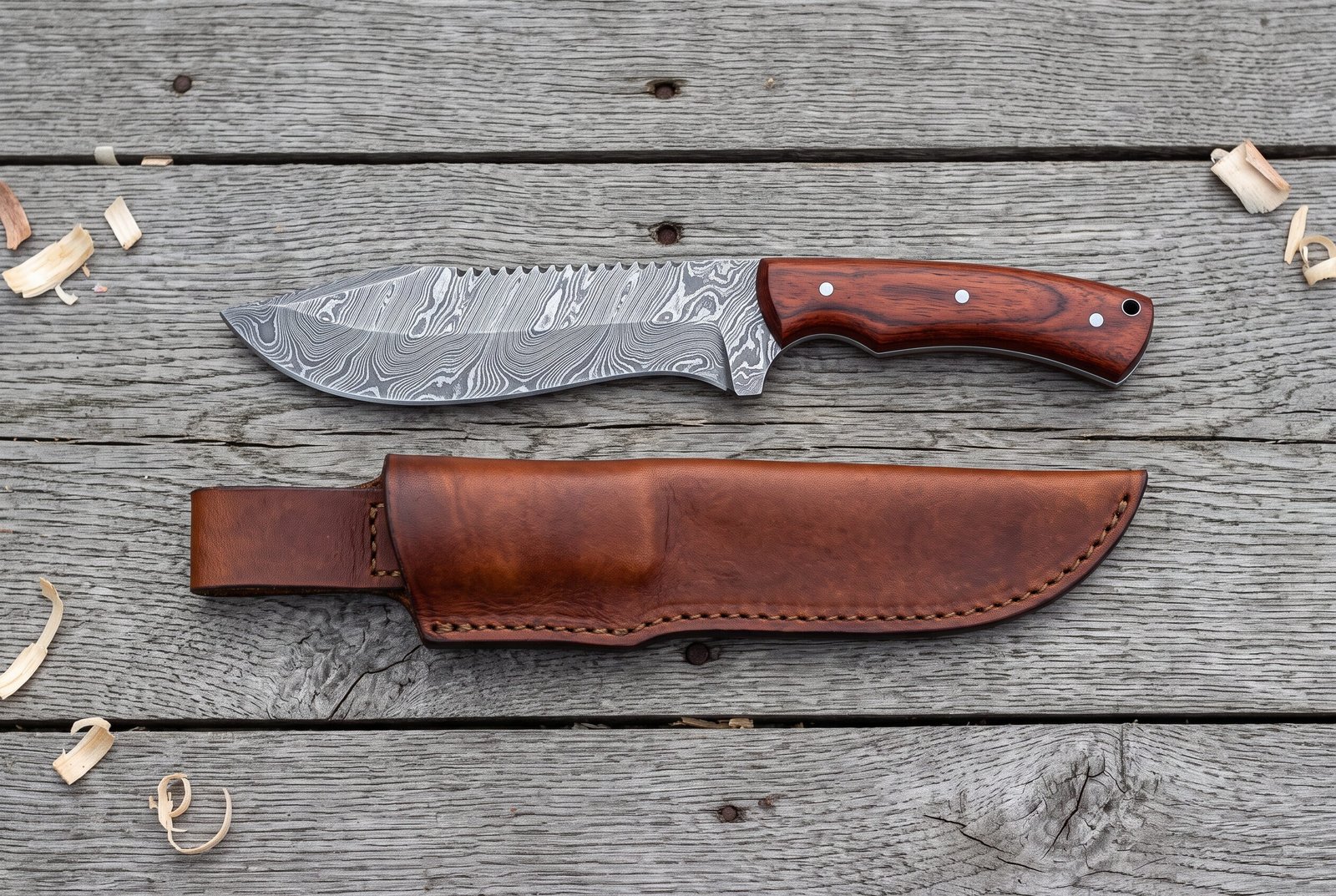 Damascus steel tracker knife with rosewood handle from above on grey boards, brown leather sheath laid below it Damascus steel tracker knife with rosewood handle from above on grey boards, brown leather sheath laid below it