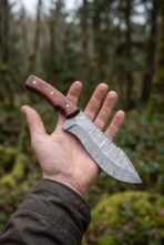 Damascus steel tracker knife with rosewood handle held across an open palm in a forest, blade pattern facing up