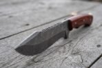 Damascus steel tracker knife in close view showing the saw toothed spine and clip point, rosewood handle behind