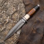 Double edged Damascus dagger with banded bone and wood handle resting on a worn brown leather hide over rock