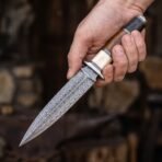 Hand gripping a Damascus dagger by its bone, wood and black handle against a blurred dark workshop background