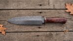 Double edged Damascus dagger with red wood handle laid flat on weathered wooden boards with oak leaves