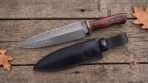 Damascus dagger with red wood handle and metal guard on rustic planks beside its black leather sheath and oak leaves