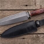 Damascus dagger with red wood handle and metal guard on rustic planks beside its black leather sheath and oak leaves