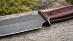 Close up of a double edged Damascus dagger blade with centre ridge, metal guard and red wood handle on a log
