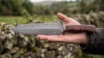 Damascus dagger with red wood handle lying across an outstretched hand in front of a dry stone wall and fields