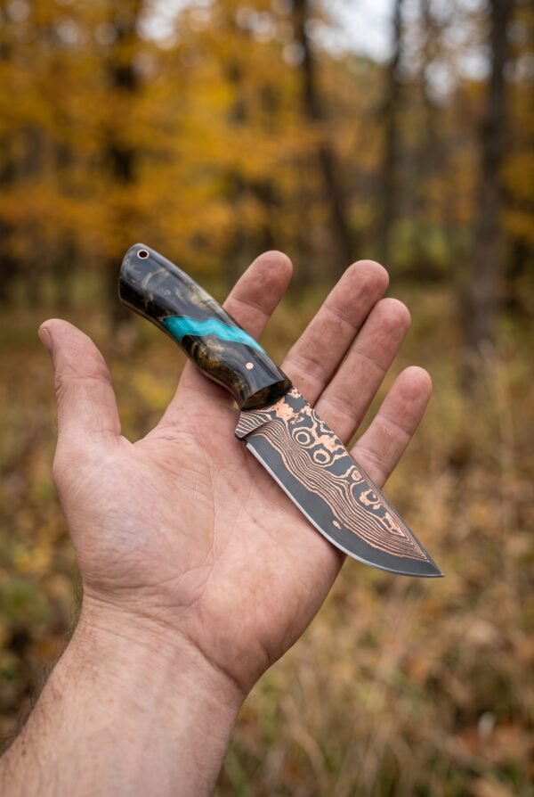 Copper Damascus skinner knife with dark and turquoise resin handle balanced on an open palm in an autumn wood