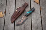 Copper Damascus skinner knife with turquoise accent resin handle lying next to a brown leather sheath on grey planks