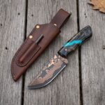 Copper Damascus skinner knife with turquoise accent resin handle lying next to a brown leather sheath on grey planks